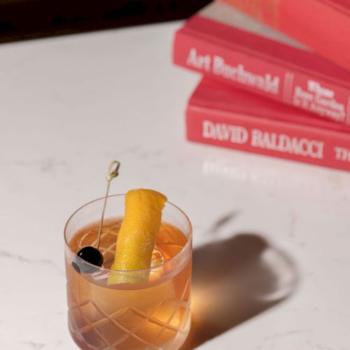 A cocktail with an orange peel and cherry sits beside stacked red books on a white surface.