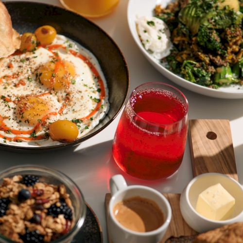 A spread with hummus, vegetables, bread, a salad, a berry granola dish, espresso, a red drink, cubes of butter, and a pastry.
