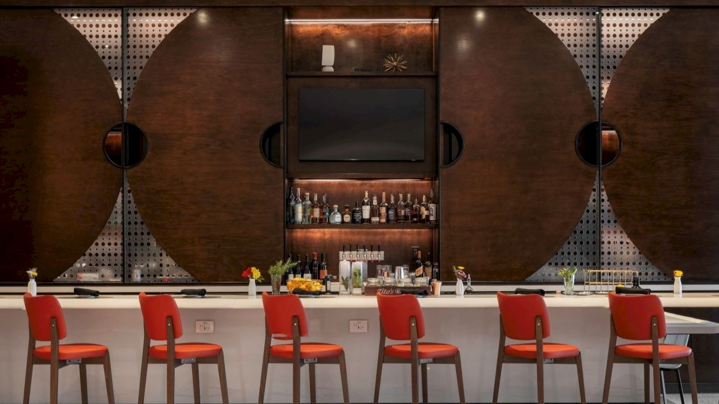 The image shows a stylish bar with red chairs, a wall-mounted TV, and a shelf stocked with bottles, set against a modern geometric backdrop.