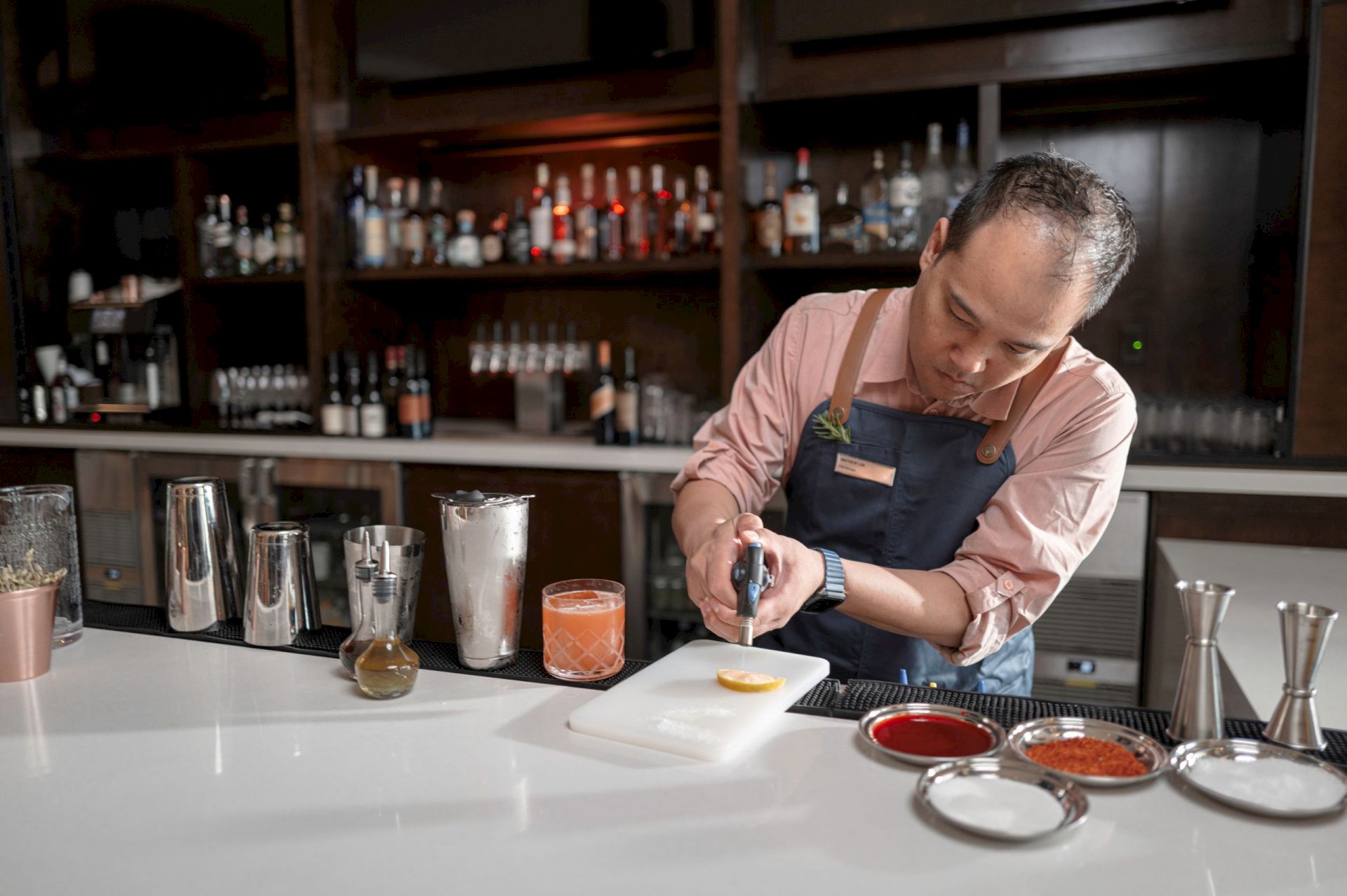A bartender prepares a cocktail, using a flame on an orange slice over a cutting board in a bar with bottles and tools.