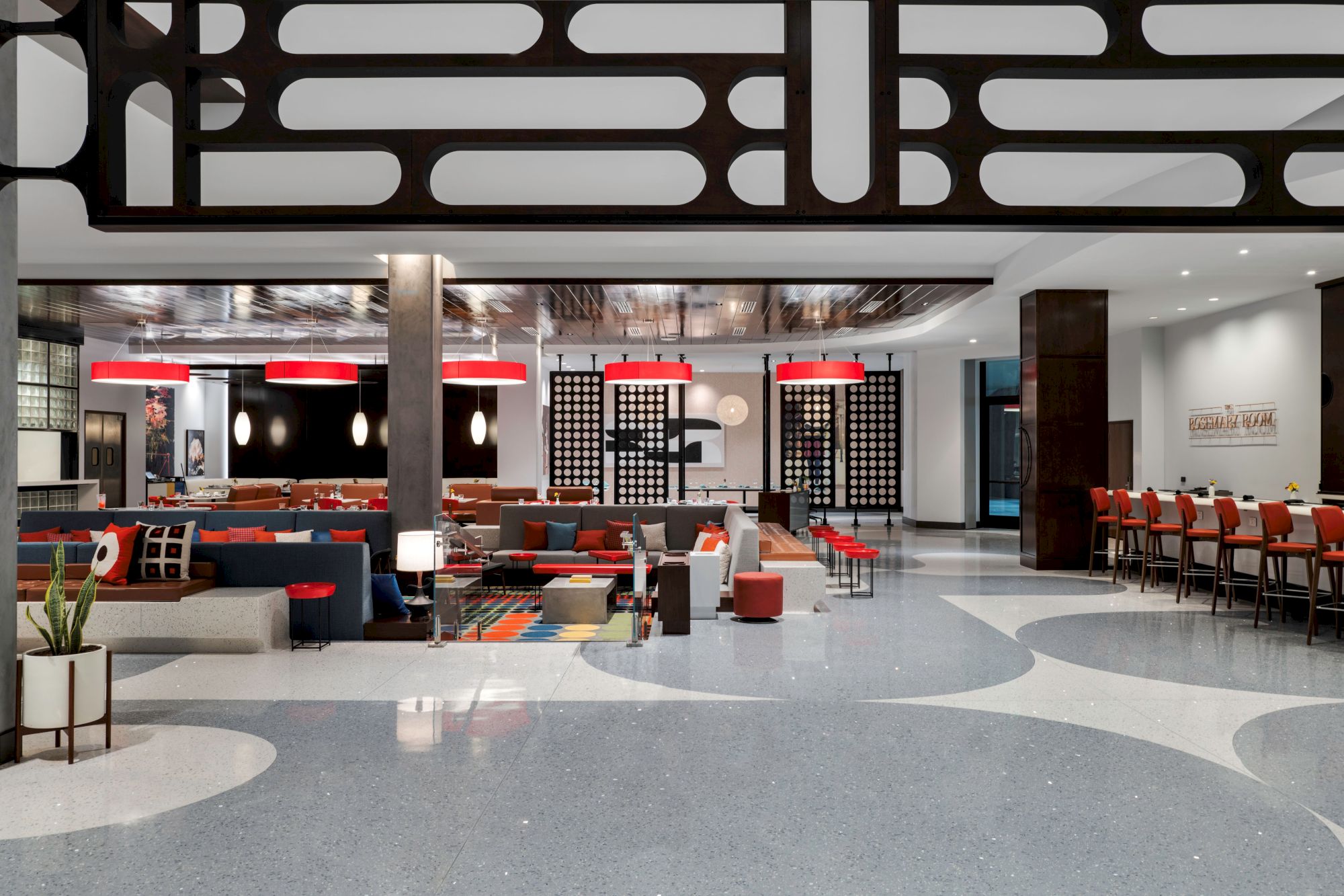 Modern office lounge with colorful sofas, red pendant lights, black panel screens, and a row of red chairs by the bar.